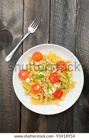 A white plate of Italian pasta fusilli with feta cheese, fresh cherry tomatoes and rucola. Served on a dark wooden rustic table, with a metal fork. Taken directly from above.