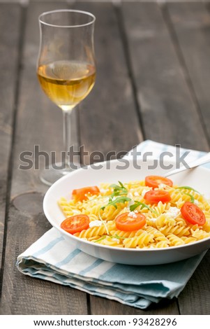 A white plate of Italian pasta fusilli with feta cheese, fresh cherry tomatoes and rucola. Served on a dark wooden rustic table, with a glass of white wine.