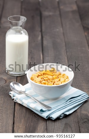 Close-up of a white bowl of cereal with a bottle of fresh milk. Served on a dark rustic wooden table, with a metal spoon on a napkin.