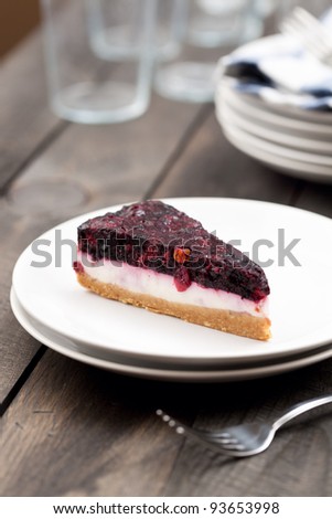 A close-up of a delicious slice of berry cheesecake on a white plate. Served on a dark wooden rustic table, with a metal fork.