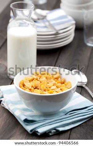 Close-up of a white bowl of cereal with a bottle of fresh milk. Served on a dark rustic wooden table, with a metal spoon on a napkin.