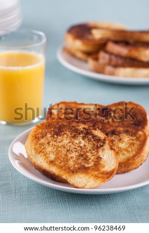 Close-up of a portion of French toast on a white plate with orange juice. Meal is served on a table with light blue tablecloth.