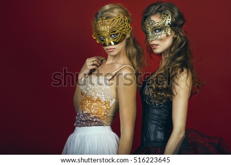 Two glam gorgeous women, blonde and brunette, in golden and bronze masks wearing evening gowns standing on red background gazing intently with expressive eyes. Studio shot. Copy-space