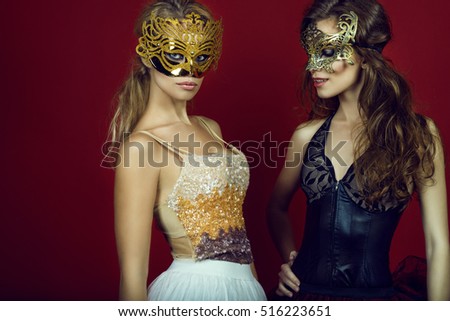 Two gorgeous young women in golden and bronze masks standing on dark red background. Blonde is staring straight, brunette looking down and smiling. Studio shot