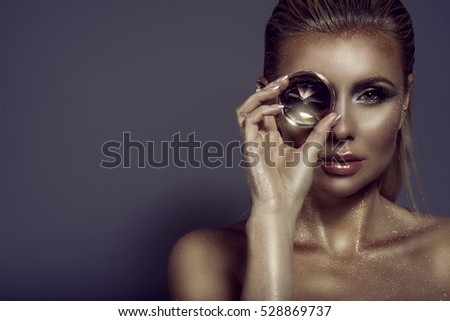 Portrait of gorgeous chic blond woman with wet hair, glittering artistic make-up and bronze skin holding a big shining gem in front of her eye. Jewellery concept. Studio shot. Close up. Copy-space