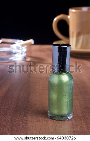 Green nail polish standing on a coffee table in front of a cup of coffee and an ashtray.