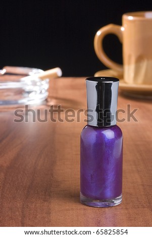 A bottle of purple nail polish standing on a coffee table in front of a cup of coffee and an ashtray.