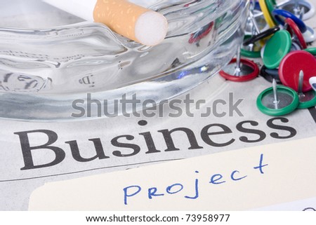 A manila project folder laying on a newspaper with the word business on it next to a glass ashtray with a filtered cigarette on it.