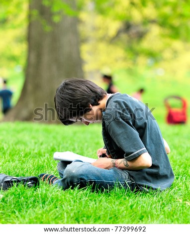 The boy the teenager holds a writing-book in a lap and draws sitting on a green grass in park.
