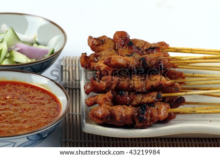 A plate of Chicken Satay served on a plate and bamboo mat with sliced onions, cucumber and a bowl of gravy