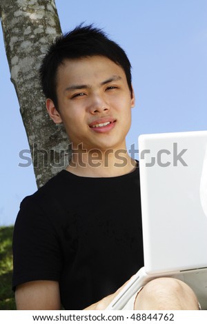 A young Asian man with a laptop leaning by a tree