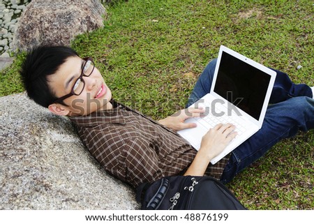 A young asian man with a laptop leaning on a rock outdoors staring upwards