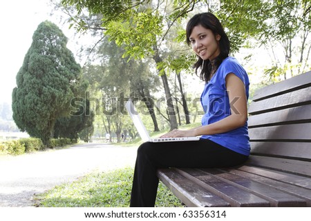 An Asian woman with a laptop sitting on a bench at a park