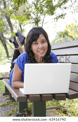 An Asian woman lying down on a bench using her laptop