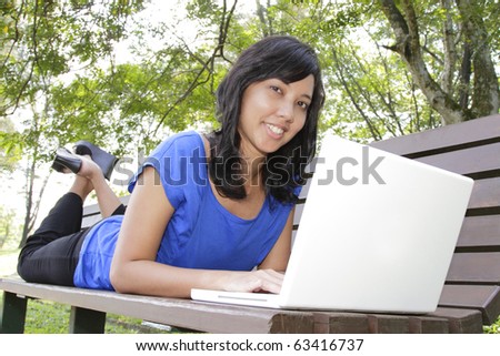 An Asian woman lying down on a bench using her laptop
