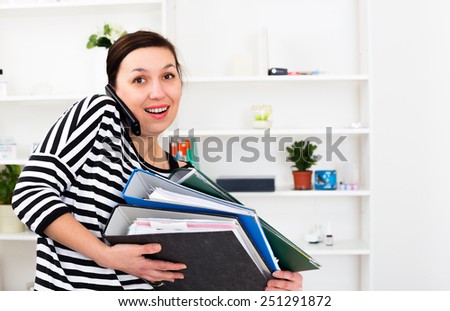 young businesswoman taking a mobile call standing balancing three large office binders on her arm