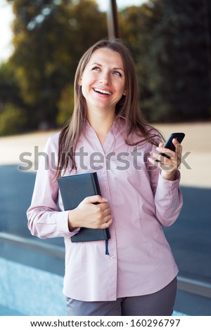 Young businesswoman with cellphone and organizer while standing against office building, outdoor