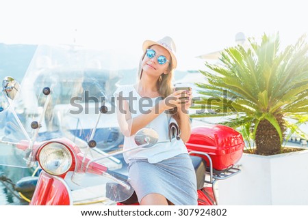 Happy young trendy woman drinking takeaway coffee near her red moped on the waterfront in the morning