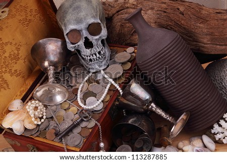A skull with coins in the eye sockets laid on a chest of coins, pearls, key and chalice.