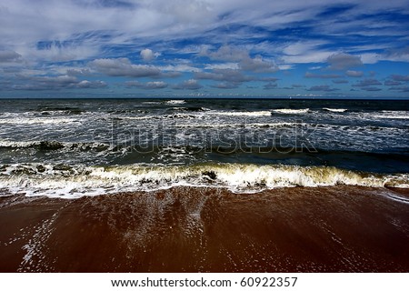 Seascape waves meeting the shore with a blue sky and white and gray clouds