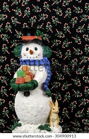 snowman figurine with rabbit holding assorted gum drops with vertical background of black cloth imprinted with candy canes