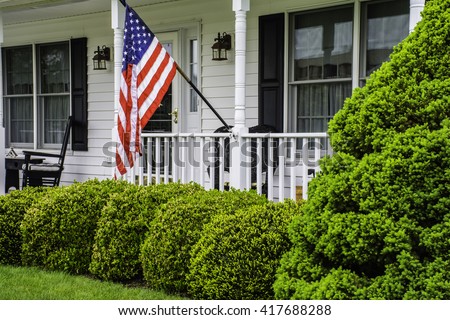 front porch of white colonial home with black shutters and rockers flying the American flag
