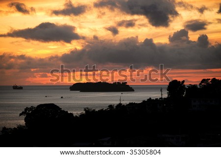 Sun setting in the west of the island of Trinidad with golden colours in the sky and think purplish blue clouds over a silhouette of the land and buildings on the coast in Cocorite