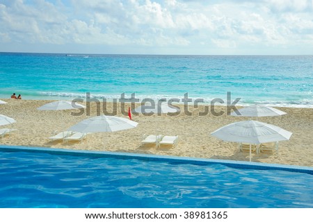 Layers of fun in the sun depicted by a diagonal of a pool white sand with white umbrellas and lounge chairs and a strip of turquoise ocean cover by a a band of blue sky with puffy cumulus cloud