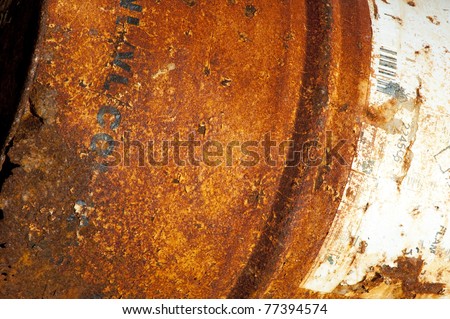 rusty and white dented metal drum with black painted markings on its surface under bright light showing stains and dents and jagged razor-sharp edges at its rim
