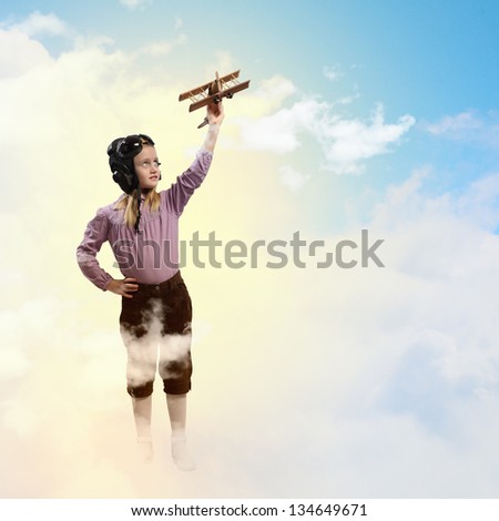 Image of little girl in pilots helmet playing with toy airplane against clouds background