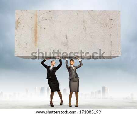 Image of two businesswomen holding stone above head. Partnership and cohesion