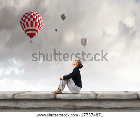 Young pretty businesswoman sitting on top of building with colorful balloons flying above