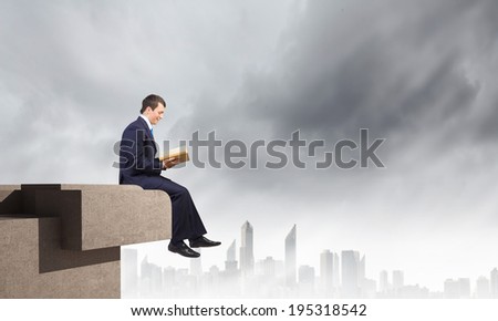 Young businessman sitting on top of building and reading book