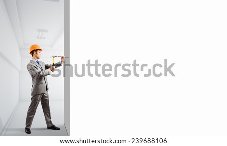 Businessman in helmet hammering nail in white wall