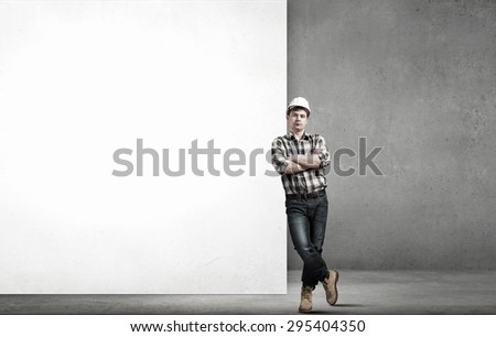 Young builder man leaning on white blank banner. Place for text