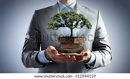 Close up of man holding pile of books with growing green tree in hands