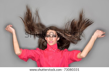 Young cute woman with long hairs flying upwards dressed in pink blouse, ring flash studio portrait on gray