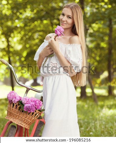 Beautiful woman wearing a nice dress having fun in park with bicycle carrying a beautiful basket full of peony flowers. Vintage scenery. Pretty girl with retro look, bike and basket with flowers