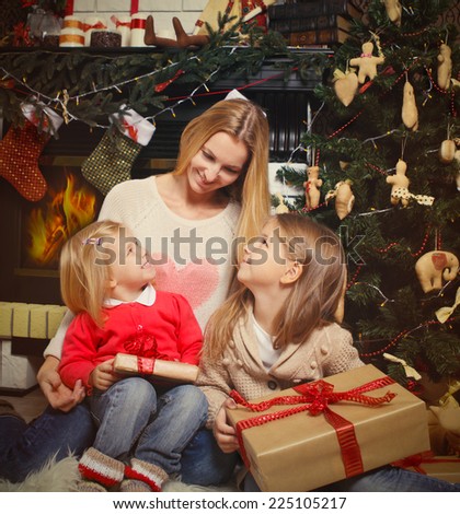 Young mother and her two little daughters with Christmas gifts by a Christmas tree in cozy living room with fireplace in winter