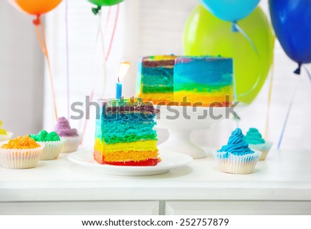 Rainbow cake and cupcakes decorated with birthday candle. Balloons on the background