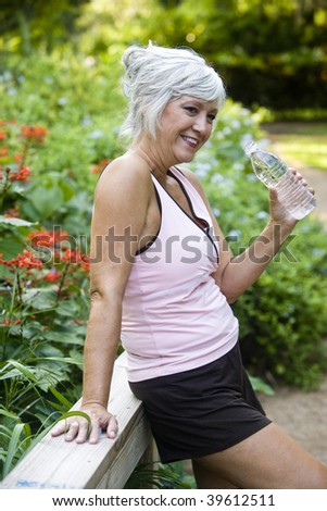 Mature woman in her 50s in workout clothes drinking a bottle of water, standing on wooden bridge in park