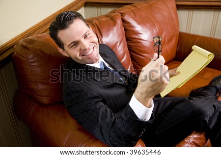 Happy businessman sitting on leather couch looking up, holding eyeglasses and notepad