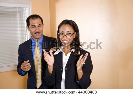 Young happy African-American female office worker standing in boardroom with middle-aged Hispanic manager smiling behind with hand on her shoulder