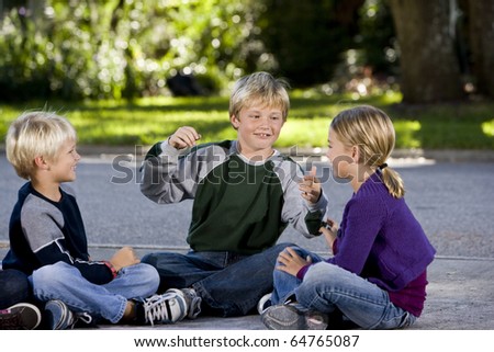 Three kids sitting on driveway playing, smiling and talking.  Ages 7 to 9