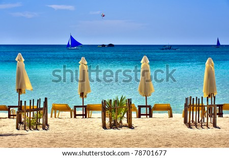 Beach Scene with Sail Boats and lounging chairs overlooking a clear blue sea.