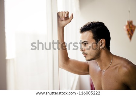 Portrait of a thoughtful handsome shirtless young man looking out of a window