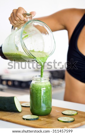 Woman pouring a green detox juice from blender to a jar, at home wearing sport clothes.