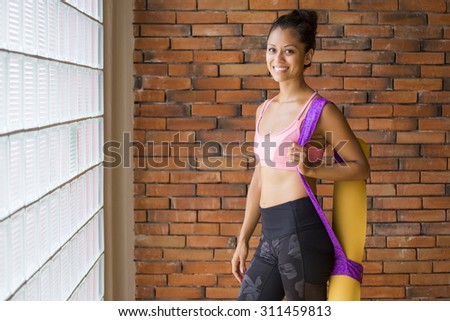 Young latin woman in a yoga studio indoors, against a brick wall, with a yoga mat.