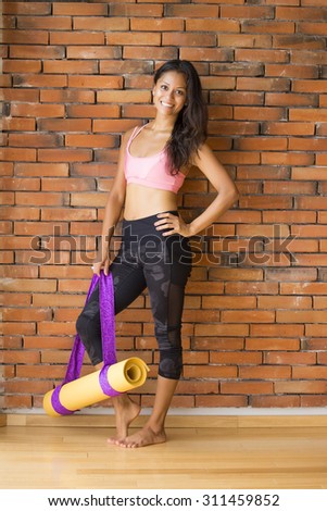 Full body shot of a latin woman in a yoga studio, against a brick wall, with a yoga mat.
