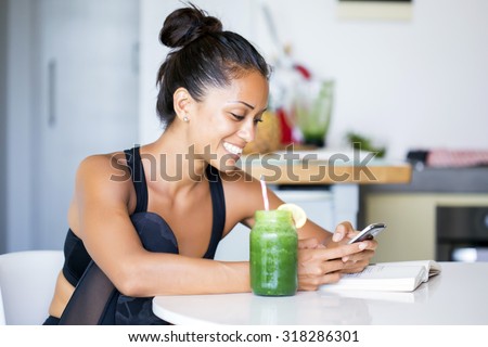 Woman drinking a homemade green detox juice, wearing sportive clothing, texting on her  phone while sitting in her kitchen table